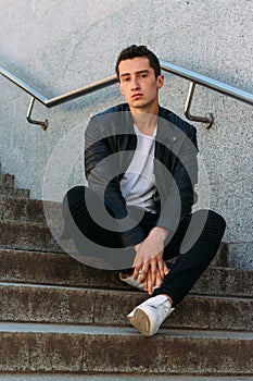 Sexy man posing sits on the steps near railing. Handsome young man in stylish black clothes and white shoes posing near a railing