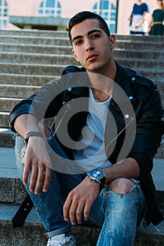Sexy man posing sits on the steps near railing. Handsome young man in stylish black clothes and white shoes watch on hand