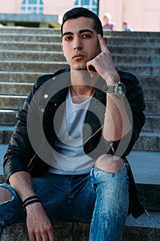 Sexy man posing sits on the steps near railing. Handsome young man in stylish black clothes and white shoes watch on hand