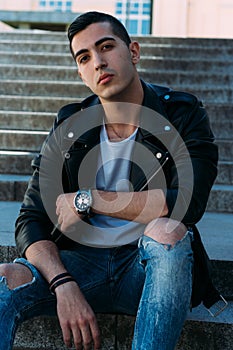 Sexy man posing sits on the steps near railing. Handsome young man in stylish black clothes and white shoes watch on hand