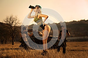 Sexy military girl with binoculars searching something ,on the highlands background with strong armed soldiers.