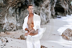 Sexy muscular man in a white shirt with a bare-chested resting on the beach, ocean waves at background.