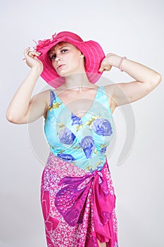 Sexy plus size girl in a blue swimsuit, fuchsia hat with brim and in a trendy bright pareo stands on a white background in the Stu