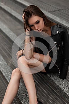 Sexy pretty fashionable young woman in in stylish autumn black clothes in leather vintage boots is resting sitting on stone steps