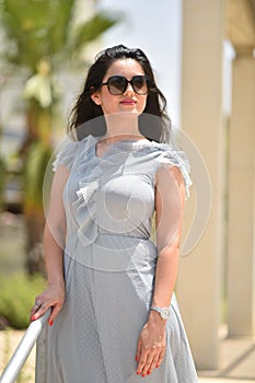 Sexy, pretty young woman on gray dress, fashion sunglasses and blue jeans stand on the street the background of a green park,