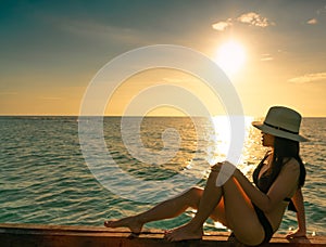 Sexy and relax woman wear black bikini with straw hat sit on wooden beam near sand beach at sunset. Girl enjoy holiday at tropical