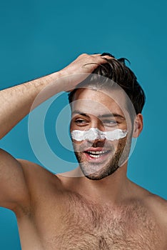 Sexy shirtless guy with white mask applied on nose for cleansing pores smiling aside, touching his brown hair isolated