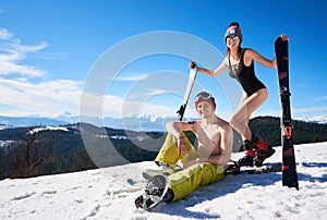 Sexy skier woman in bikini and snowboarder man with bare torso on background of snowy mountains.