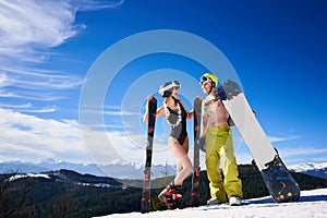 Sexy skier woman in bikini and snowboarder man with bare torso on background of snowy mountains.