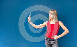 Sexy slim blonde womanin red t-shirt on a blue studio background making selfie with smartphone. Copyspace