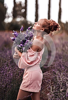 Sexy slim red-haired woman in jumpsuit with shorts stands in the middle of field holding bouquet of wildflowers lavender