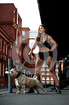 Sexy and srong brunette woman posing with bully dog on city street, looking at camera.