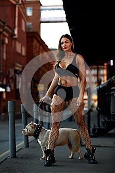 Sexy and srong brunette woman posing with bully dog on city street, looking at camera.