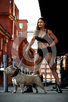 Sexy and srong brunette woman posing with bully dog on city street, looking at camera.