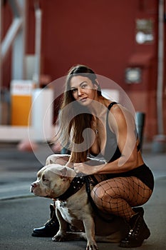 Sexy and srong brunette woman posing with bully dog on city street, looking at camera.