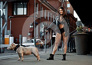 Sexy and srong brunette woman posing with bully dog on city street, looking at camera.