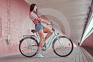 stylish brunette girl wearing a pink flannel shirt and denim shorts in glasses, posing with city bike against a