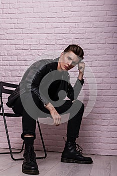 Sexy stylish model of a young man in fashionable black clothing in trendy leather shoes is resting on a chair near a vintage brick