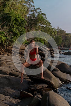 Sexy tanned brunette in red bikini sitting on the stone among tropical beach