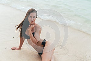 tanned girl in black swimsuit posing on sandy beach near ocean. Beautiful model sunbathes and rests