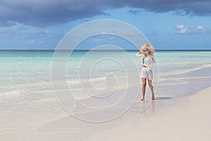 Sexy tanned girl in blue bikini and white tunica running on the seashore. Beautiful model sunbathes and rests on sea. Concept