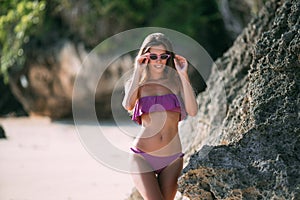 Sexy tanned girl in purple swimsuit posing on beach with sand and large stones