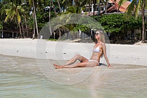 Sexy tanned girl in white swimsuit posing on sandy beach. Beautiful model sunbathes and rests on ocean shore. Concept vacation,