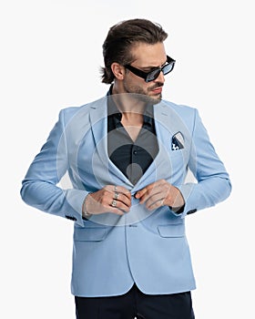 sexy unshaved guy with sunglasses looking to side while fixing suit