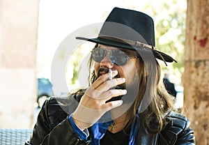 white man with sunglasses and a fedora hat smoking a cigarette.