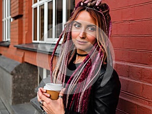 Sexy woman with big brown eyes and hairstyle with pink dreadlocks and pigtails in black clothes is holding coffee. Shot outdoors
