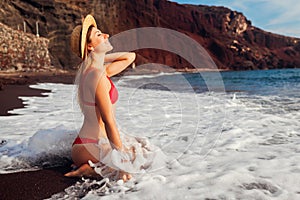 Sexy woman in bikini relaxing on Red beach in Santorini, Greece. Girl sitting on sand. Summer vacation trip
