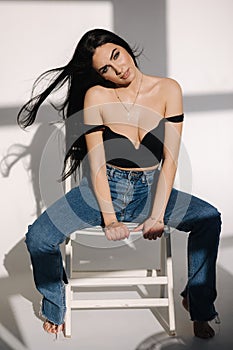 Sexy woman in black topic and blue jeans sits on chair during studio photoshoots. Brunette girl posing to photographer