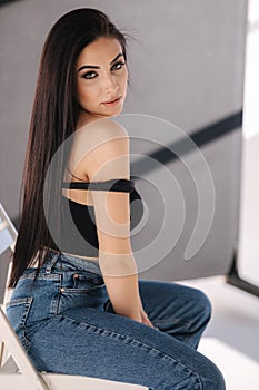 Sexy woman in black topic and blue jeans sits on chair during studio photoshoots. Brunette girl posing to photographer