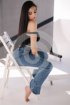 Sexy woman in black topic and blue jeans sits on chair during studio photoshoots. Brunette girl posing to photographer