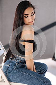 Sexy woman in black topic and blue jeans sits on chair during studio photoshoots. Brunette girl posing to photographer