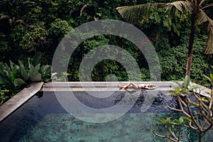 Slim brunette woman in swimsuit relaxing on edge infinity pool in jungle. Palms around and crystal clean water