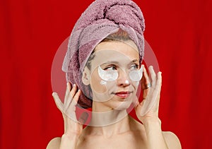 Sexy woman posing on red background. She does beauty treatments in a towel on her head. Patches under the eyes. Hands near the