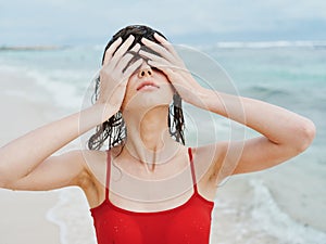 Sexy woman in red swimsuit on ocean beach with wet hair covers face with hands, sunscreen