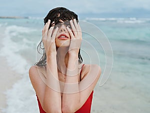 Sexy woman in red swimsuit on ocean beach with wet hair covers face with hands, sunscreen