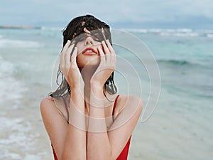 Sexy woman in red swimsuit on ocean beach with wet hair covers face with hands, sunscreen