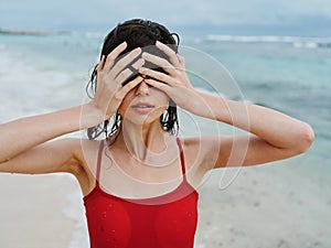 Sexy woman in red swimsuit on ocean beach with wet hair covers face with hands, sunscreen