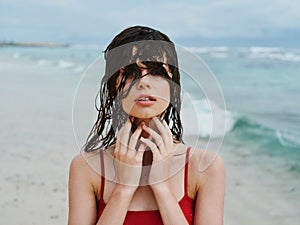 Sexy woman in red swimsuit on ocean beach with wet hair covers face with hands, sunscreen