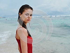 Sexy woman in red swimsuit on ocean beach with wet hair looking at camera, slim pumped body sunscreen