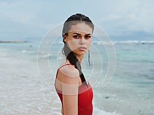 Sexy woman in red swimsuit on ocean beach with wet hair looking at camera, slim pumped body sunscreen