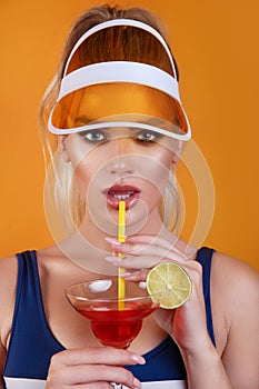 Sexy woman in swimsuit and trendy plastic visor holding glass of fresh drink on yellow background