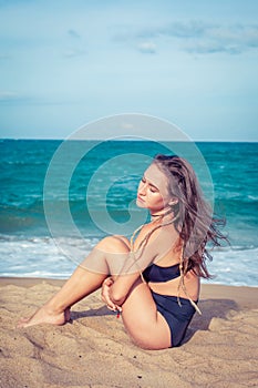 woman with tan skin in a black swimsuit sitting on the sand of the beach. Female hug her knees near sea.