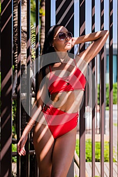 woman wearing red bikini swimsuit and sunglasses standing on the near swimming pool with artistic shadow pattern on