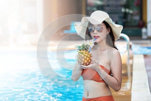 Sexy young asian woman in swimsuits in summer. beautiful woman in orange bikini with hat sitting in swimming pool with tropical