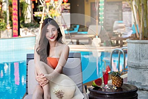 Sexy young asian woman in swimsuits in summer. beautiful woman in orange bikini with tropical drink sitting on sofa by swimming