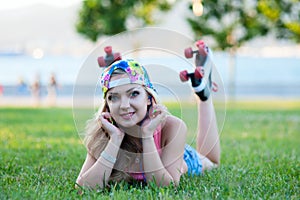 young blond girl in vintage rollers lying on lawn in park and listening to music on headphones